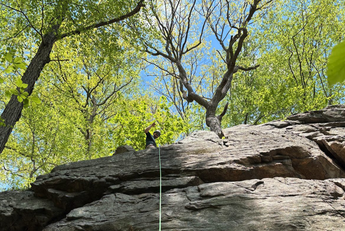 Eliot waving from the top of a rock climbing route, under spring trees