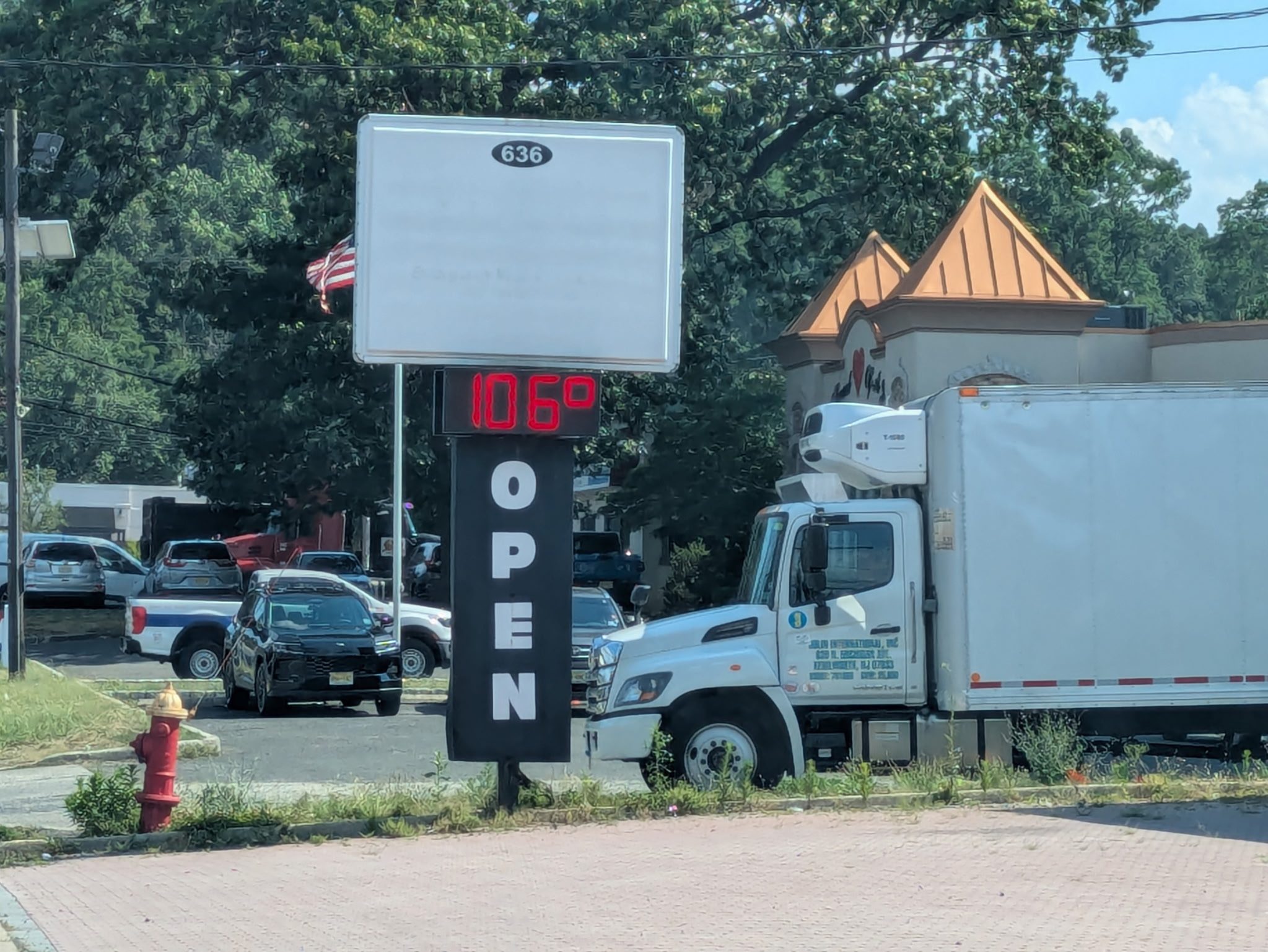 A roadside business sign on North Michigan Avenue in Kenilworth with a red digital thermometer reading 106 degrees above a tall OPEN sign, trees and a delivery truck behind it.